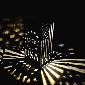 May include: A black metal lantern with cutouts that project light in the shape of stars and stripes. The design features the text 1776 surrounded by a circle of stars, the letters USA, and a vertical American flag pattern on the side panels.