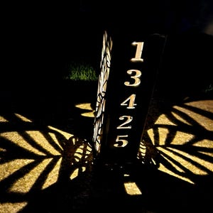 May include: A vertical rectangular house number sign featuring the digits 13425 cut out from a dark metal panel. The sign is illuminated from within, casting bright light through the numbers and a decorative leaf pattern, creating shadows.