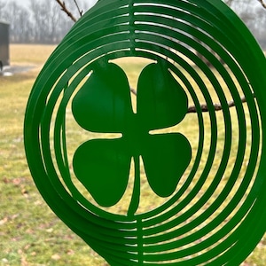 May include: A green metal wind spinner with a four-leaf clover design. The spinner is made of multiple layers of metal that are cut into a circle shape. The clover is in the center of the spinner and is also made of metal.