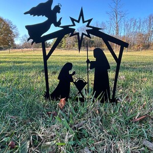 May include: Black metal silhouette of a nativity scene with a star, angel, Mary, Joseph, and a manger. The scene is set against a green grassy background.