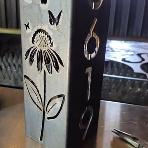 May include: A rectangular metal house number sign featuring the laser-cut numbers 6019 vertically aligned on the side. The front panel displays a cut-out design of a single flower with a stem and leaves, surrounded by several small butterflies.