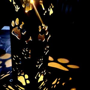 May include: A black rectangular lantern featuring cutouts of paw prints and bones. The interior light shines through these openings, casting golden paw print and bone-shaped shadows onto the surrounding area, creating a glowing effect.
