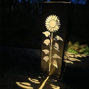 May include: A rectangular black metal solar lantern featuring a laser-cut sunflower design on its front panel. The internal light source illuminates the cutout, casting a bright, detailed sunflower shadow pattern onto the ground in front of the unit.