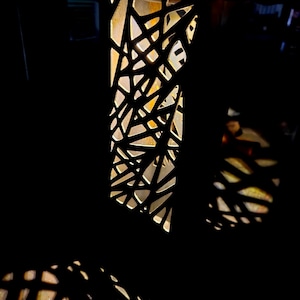 May include: A rectangular lamp with a dark, laser-cut outer shell featuring an intricate, irregular geometric lattice pattern. The interior emits a warm, soft golden light that shines through the open spaces of the dark frame, casting shadows.