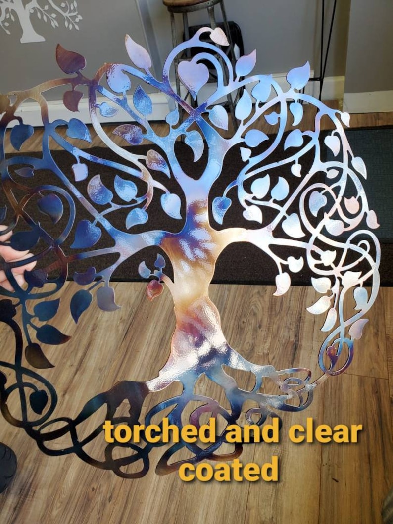 Op de afbeelding: Een metalen wandkunstwerk in de vorm van een levensboom met een blauwe, paarse en oranje iriserende afwerking. De boom heeft een gedetailleerd ontwerp met ingewikkelde takken en bladeren. De tekst "torched and clear coated" is zichtbaar onderaan de afbeelding.