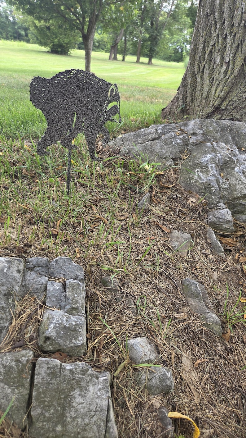 May include: A black metal silhouette of a raccoon standing on a thin stake. The raccoon figure features a detailed, textured pattern across its body, capturing the animal in a side profile stance with its head turned slightly downward.