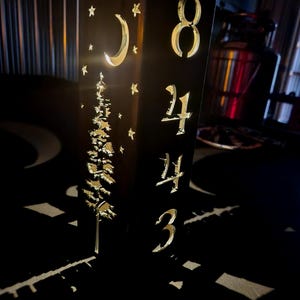 May include: A dark wooden address sign with gold-colored numbers and decorative cutouts. The sign features the numbers 8443, a crescent moon, stars, and a tree design. The sign is illuminated from within, creating a warm glow.