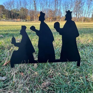 May include: Three black metal silhouettes of the three wise men, walking in a line, holding gifts. The silhouettes are standing on a green grassy surface.