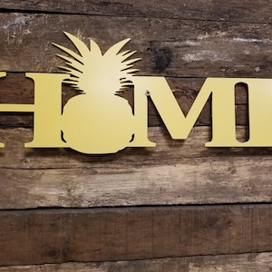 May include: Gold metal home sign with a pineapple silhouette in the center. The sign is mounted on a rustic wooden background.