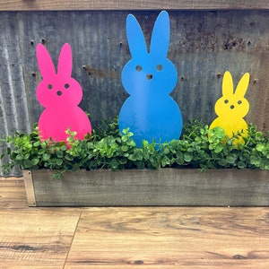 May include: Three Easter bunny shaped metal cutouts in a wooden planter. The bunnies are pink, blue, and yellow. The planter is filled with green artificial greenery.