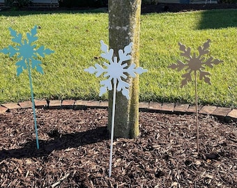 May include: Three metal snowflake yard decorations in different colors. The snowflakes are white, light blue, and gold. They are all on stakes and are placed in a bed of brown mulch.