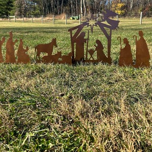 May include: A metal nativity scene silhouette with a star, a manger, a donkey, a cow, and figures of Mary, Joseph, and the baby Jesus. The figures are all brown and are standing on a green grassy lawn.