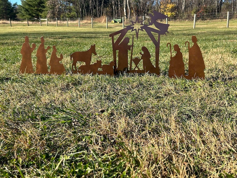 May include: A metal nativity scene silhouette with a star, a manger, a donkey, a cow, and figures of Mary, Joseph, and the baby Jesus. The figures are all brown and are standing on a green grassy lawn.