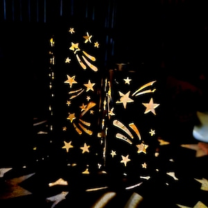 May include: Two black metal lanterns with star and shooting star cutouts. The lanterns are illuminated from within, casting star-shaped shadows. The design creates a celestial theme, suitable for home decor or special events.