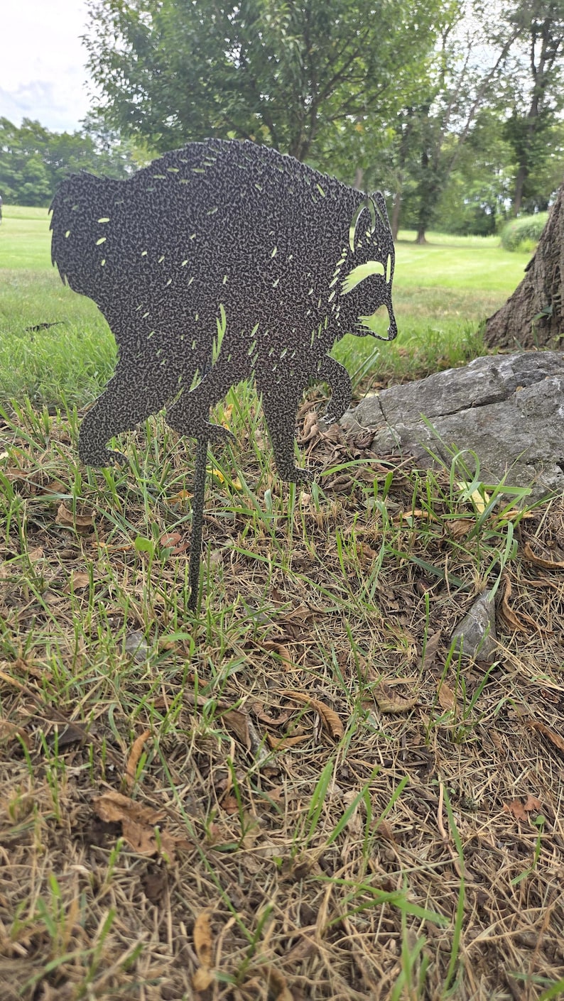 May include: A dark gray, textured metal garden stake shaped like a raccoon in a walking pose. The silhouette features cut-out details for the eyes, ears, and fur texture, mounted on a thin metal rod for insertion into the ground.