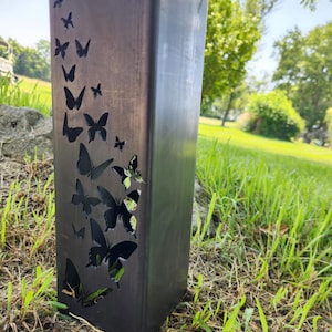 May include: A dark metal outdoor pillar with a rectangular shape. The pillar features a series of butterfly cutouts along one side. The pillar is set on a grassy area with a blurred background of trees and sky.