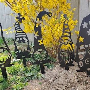 May include: Five black metal silhouette gnomes with different hats and facial features. The gnomes are standing in a row in front of a bush with yellow flowers.