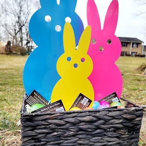 May include: Three metal bunny rabbits in a black wicker basket. The bunnies are blue, yellow, and pink. The basket is filled with colorful Easter eggs and business cards for Rhodes Welding & Metal Works.