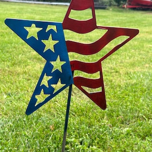 May include: A metal star garden stake with a patriotic design. The star features a blue section with cut-out stars and a red section with stripes, resembling the American flag. The stake is placed in green grass.