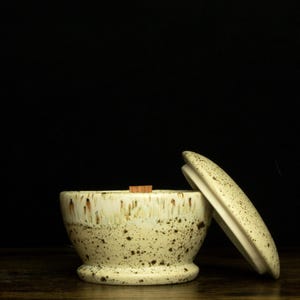 May include: A speckled ceramic bowl with a lid, set against a dark background. The bowl is off-white with brown speckles and a wooden piece inside. The lid is slightly ajar, revealing the interior.