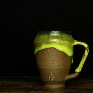 Könnte beinhalten: Eine braune Keramiktasse mit grüner und gelber Glasur. Die Tasse hat einen abgerundeten Körper und einen Henkel. Die Tasse hat ein kleines, grünes Blattdesign auf der Vorderseite.