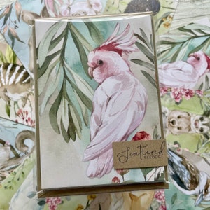 May include: A greeting card featuring a watercolor illustration of a pink and white cockatoo with a red crest, perched among green leaves. The card has a brown tag that reads "Scattered Seedco."