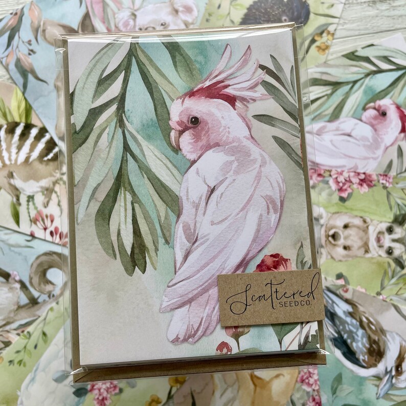 May include: A greeting card featuring a watercolor illustration of a pink and white cockatoo with a red crest, perched among green leaves. The card has a brown tag that reads "Scattered Seedco."