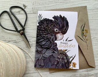 Op de afbeelding: Een felicitatiekaart met een zwarte kaketoe met gouden stippen op zijn veren. De vogel is in profiel afgebeeld en kijkt naar links. De kaart heeft een witte achtergrond en de tekst "Happy Birthday" is geschreven in zwarte cursieve letters.