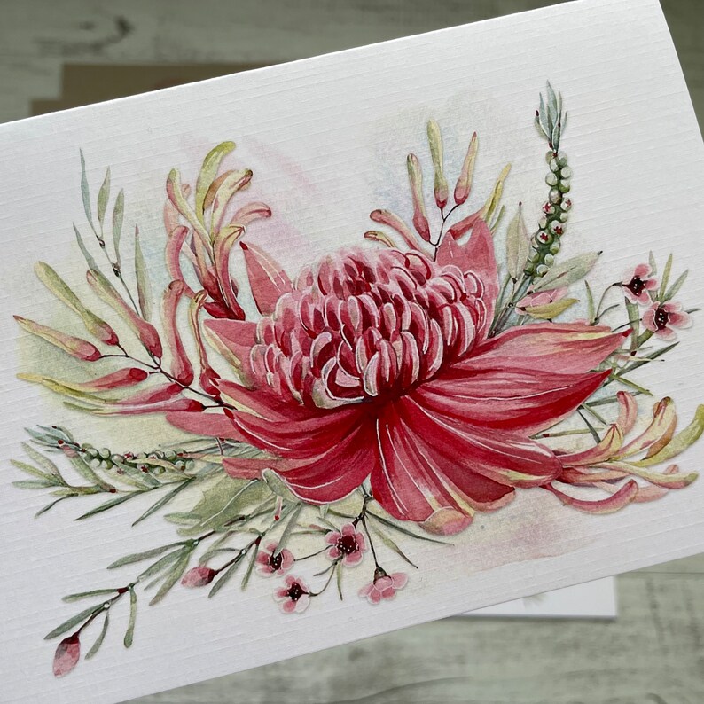 Op de afbeelding: Een aquarel schilderij van een roze bloem met groene bladeren en stengels. De bloem heeft een groot, rond midden en bloemblaadjes die naar buiten gekruld zijn. De bladeren zijn in verschillende tinten groen en zijn rond de bloem in een los, vloeiend patroon gerangschikt.