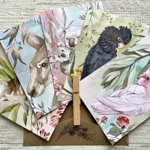 May include: A set of watercolor greeting cards featuring Australian wildlife. The cards depict a bilby, kangaroo, koalas, a black cockatoo, and a pink cockatoo. The cards are arranged with a brown envelope and a wooden clothespin.
