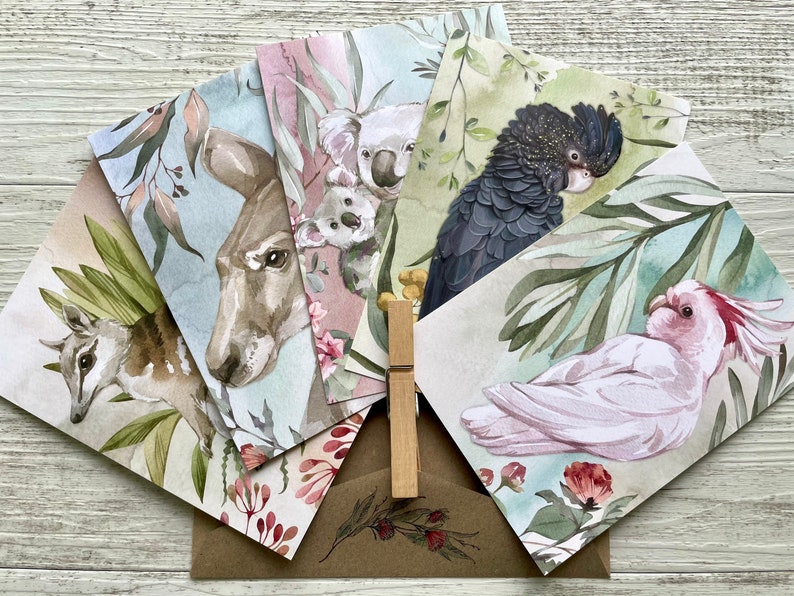 May include: A set of watercolor greeting cards featuring Australian wildlife. The cards depict a bilby, kangaroo, koalas, a black cockatoo, and a pink cockatoo. The cards are arranged with a brown envelope and a wooden clothespin.
