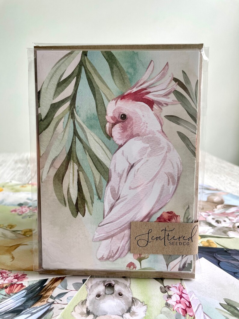 May include: A set of greeting cards featuring a watercolor illustration of a pink and white cockatoo with a red crest, set against a backdrop of green leaves. The cards are packaged in a clear plastic sleeve, with the text "Scattered Seedco" on a brown label.