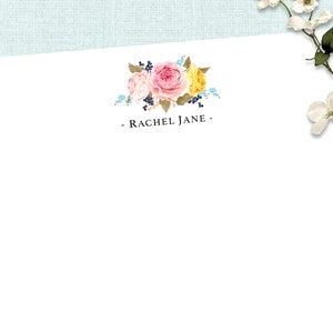Op de afbeelding: Een witte briefpapierkaart met een bloemmotief met roze en gele rozen, blauwe bessen en groene bladeren. De tekst "RACHEL JANE" is in zwart onderaan de kaart gedrukt.