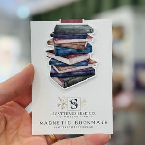 May include: A magnetic bookmark with a watercolour illustration of a pile of books in shades of blue, brown, and green. The bookmark displays the text "MAGNETIC BOOKMARK" and the brand name "SCATTERED SEED CO."