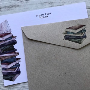 Puede incluir: Una tarjeta blanca con el texto "A Note From Jonah" en la parte superior. La tarjeta presenta una ilustraci&oacute;n de acuarela de una pila de libros. La tarjeta est&aacute; junto a un sobre marr&oacute;n con una ilustraci&oacute;n de acuarela de una pila de libros m&aacute;s peque&ntilde;a.