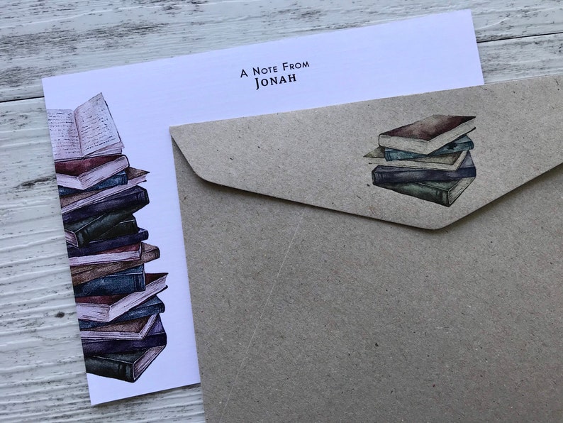 Puede incluir: Una tarjeta blanca con el texto "A Note From Jonah" en la parte superior. La tarjeta presenta una ilustraci&oacute;n de acuarela de una pila de libros. La tarjeta est&aacute; junto a un sobre marr&oacute;n con una ilustraci&oacute;n de acuarela de una pila de libros m&aacute;s peque&ntilde;a.