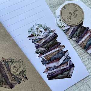 May include: A white envelope with a stack of books and flowers on the front. The envelope has a brown paper flap and a brown paper backing. The books are stacked in a pile and have a watercolor effect. The flowers are white and green and are arranged in a bouquet. The envelope has a small brown tag with the text "Lettered Treasures" on it.
