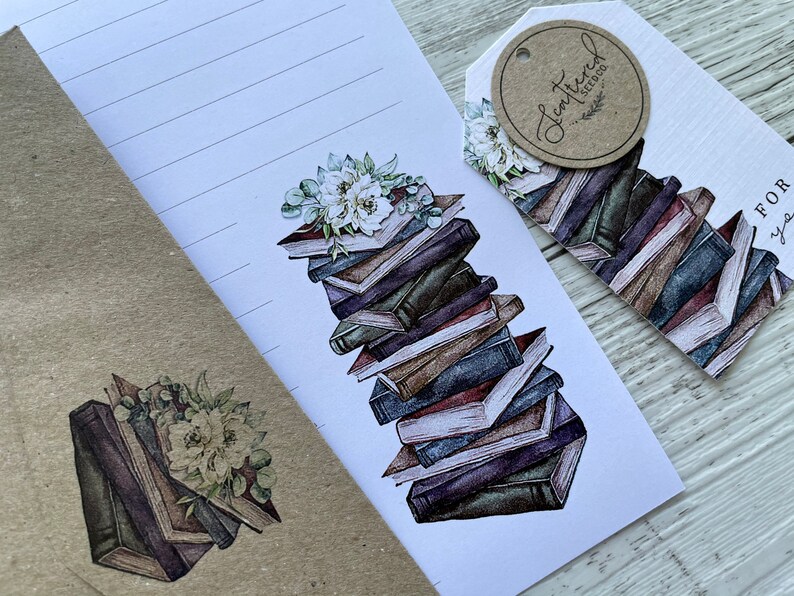 May include: A white envelope with a stack of books and flowers on the front. The envelope has a brown paper flap and a brown paper backing. The books are stacked in a pile and have a watercolor effect. The flowers are white and green and are arranged in a bouquet. The envelope has a small brown tag with the text "Lettered Treasures" on it.