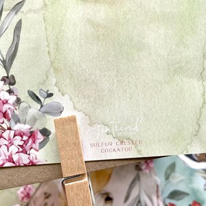 May include: A watercolor print featuring a floral arrangement with pink and gray flowers and green foliage. The print has a light green and beige background. The text "Shattered Seed Co" and "SULFUR CRESTED COCKATOO" are visible.