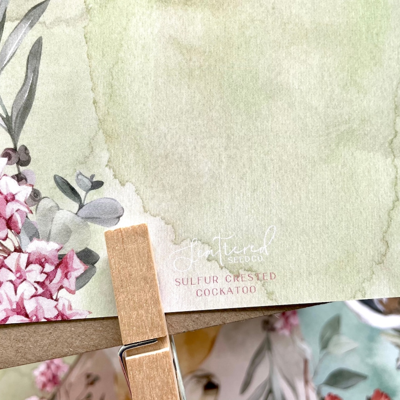 May include: A watercolor print featuring a floral arrangement with pink and gray flowers and green foliage. The print has a light green and beige background. The text "Shattered Seed Co" and "SULFUR CRESTED COCKATOO" are visible.