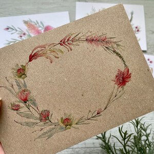 Op de afbeelding: Een bruine kaart met een aquarelkrans van roze en rode bloemen en groene bladeren. De krans is ovaalvormig en bevindt zich in het midden van de kaart.