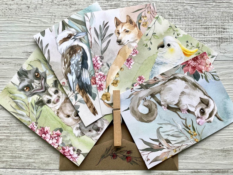 May include: A collection of watercolor greeting cards featuring Australian animals: a kookaburra, dingo, cockatoo, sugar glider, and others. Each card showcases the animal amidst floral and foliage elements, with a kraft paper envelope and wooden clip.