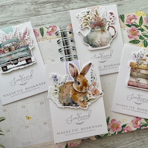 May include: Four watercolour-style magnetic bookmarks with floral designs. The bookmarks feature a teapot with flowers, a truck with flowers, a rabbit in a field of flowers, and a stack of books with flowers. Each bookmark has the text "Scattered Seedco" and "Magnetic Bookmark" with a website address.