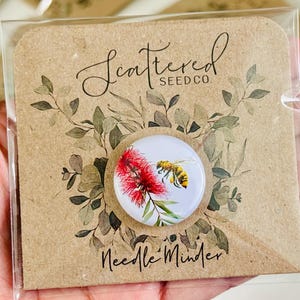 May include: A round needle minder featuring a bee and red flower design. The needle minder is on a kraft paper backing with the text "Scattered Seed Co." and "Needle Minder". The background has a floral design.