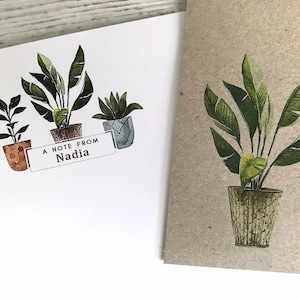May include: A white card with a handwritten message that reads "A note from Nadia" and three potted plants. The plants are drawn in a watercolor style with green leaves and brown pots. The card is on a white wooden surface.