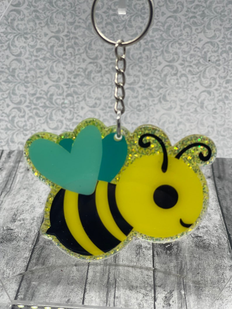 May include: A yellow and black bee keychain with a teal heart and glitter accents. The bee has a smiling face and black eyes.