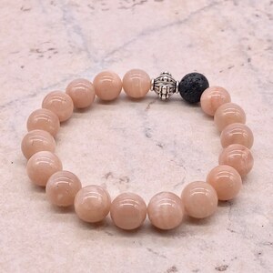 May include: A bracelet made with light pink sunstone beads and a black lava bead. The bracelet has a silver bead with a decorative design.