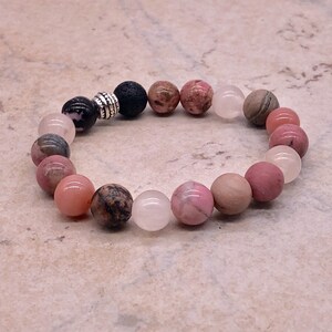 May include: A pink, gray, and black beaded bracelet with a silver spacer bead. The bracelet is made with natural stones and is designed to be worn on the wrist.