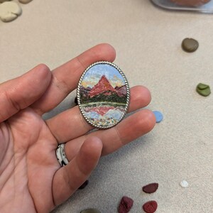 K&ouml;nnte beinhalten: Ovaler Anh&auml;nger mit einem Berglandschaftsgem&auml;lde. Das Kunstwerk zeigt einen roten Berg, gr&uuml;ne B&auml;ume und einen reflektierenden See unter blauem Himmel. Der Anh&auml;nger hat einen silberfarbenen, gedrehten Seilrand.