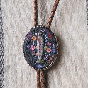 May include: A bolo tie featuring an oval-shaped pendant with a dark gray background. The pendant displays a central rainbow trout surrounded by colorful flowers, green leaves, and small white stars. The pendant has a silver-toned twisted metal border.